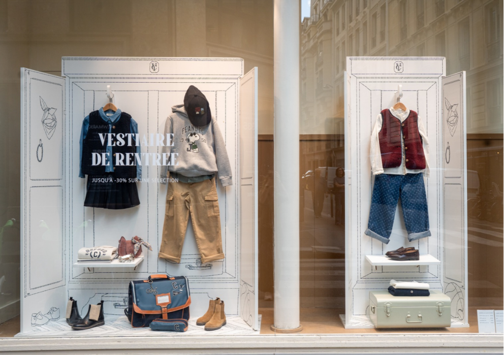 Vitrine Cyrillus de rentrée avec deux armoires blanches illustrées au trait noir, vêtements enfant suspendus sans mannequin, cartable et malle, scénographie par Twist Design